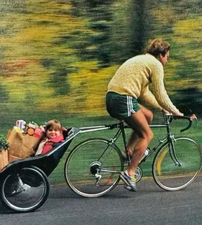A cyclist in a cable-knit sweater towing a smiling child and a load of groceries.