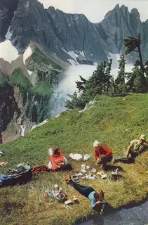 Hikers picnicking in a mountain meadow, one drinking straight from a stream.
