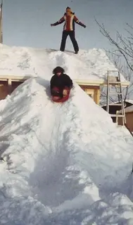 A kid sledding down a snowbank piled as high as the roof of a house.